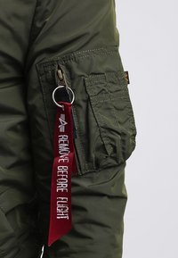 Manica di giacca verde con tasca con zip e etichetta di stoffa rossa attaccata che recita "RIMUOVERE PRIMA DEL VOLO" su un anello metallico.