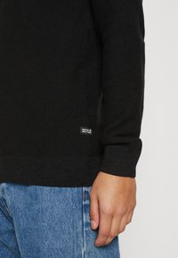 Svart strikkegenser med ribbestruktur og rund hals, med et lite merke på siden. Kombinert med lyseblå denimjeans.