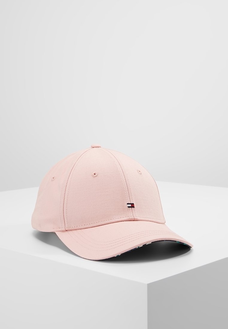 Casquette de baseball rose clair avec petit logo rectangulaire sur le devant, reposant sur une surface blanche sur fond gris clair uni.