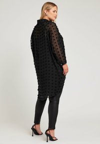 Svart genomskinlig långärmad topp med texturerade prickar, krage, sidofickor och en något längre bakdel; kombinerad med åtsittande svarta läderleggings.