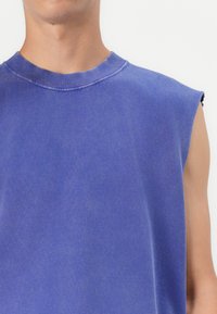 Ärmelloses blaues T-Shirt aus weicher Baumwolle. Verfügt über einen runden Halsausschnitt und rohe Saumkanten, mit einem leicht verblassten, strukturierten Aussehen.