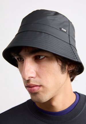 Jeune homme portant un chapeau bob imperméable noir avec une étiquette « RAINS » et un t-shirt col rond sombre sur un fond clair.