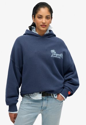 Marineblauwe hoodie met een losse pasvorm, voorzien van een palmboomgraphic en logo op de borst, gecombineerd met lichtblauwe denim.