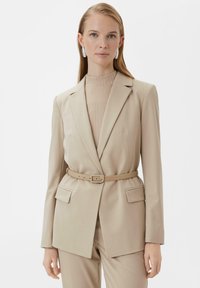 Beżowa marynarka o dopasowanym kroju z paskiem w talii, dwoma przednimi kieszeniami i szpiczastymi klapami. Teksturowany beżowy golf pod spodem dopełnia całość.