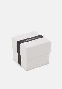 Caja de regalo de cartón blanco con una banda negra que presenta el logo de "Calvin Klein", de forma cuadrada, textura suave y diseño minimalista.