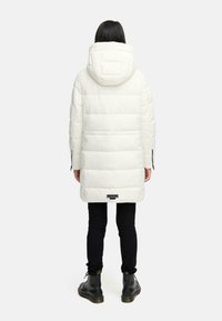 Vit puffjacka med huva, med horisontell quiltad design, sidodragkedjor och en etikett vid bakre fållen. Kombinerad med svarta byxor och stövlar.