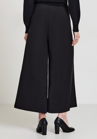 Pantaloni neri a gamba larga in tessuto liscio, con vita alta, pieghe e orlo senza cuciture. Abbinati a tacchi neri.