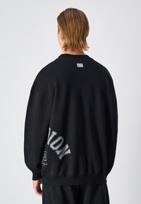 Sweatshirt noir avec col rond, coupe oversized et texte graphique argenté au dos. Fabriqué en tissu doux avec des poignets côtelés.