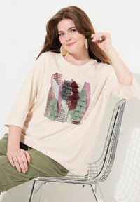 Cremfarbener T-Shirt mit 3/4-Ärmeln, das mit einem strukturierten Stoffapplikation in Grün, Rot und Grau versehen ist. Getragen mit olivgrünen Hosen, sitzend auf einem Drahtstuhl.