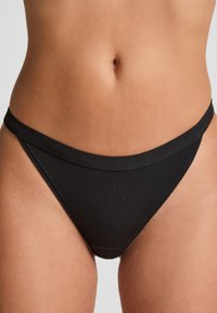 Milieu de taille d'une femme portant des sous-vêtements de style bikini noir uni sur un fond neutre.