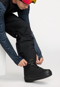 Botte de snowboard noire avec un dessus texturé et une semelle en caoutchouc, en train d'être attachée par une personne portant une chemise à manches longues à motifs et un pantalon.