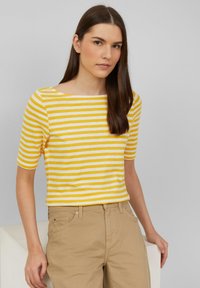 Haut jaune et blanc à rayures, à manches courtes et au col bateau ; textile doux ; associé à un pantalon beige, sur un fond neutre.