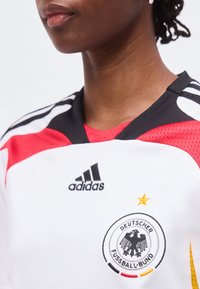 Personne portant un maillot de football allemand blanc et rouge avec le logo Adidas et l'emblème du Deutscher Fussball-Bund représentant un aigle noir et une étoile dorée.