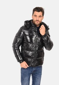 Giubbotto imbottito nero con finitura lucida, chiusura con zip frontale, cappuccio regolabile e polsini elasticizzati. Indossato sopra un maglione blu navy e jeans di denim.
