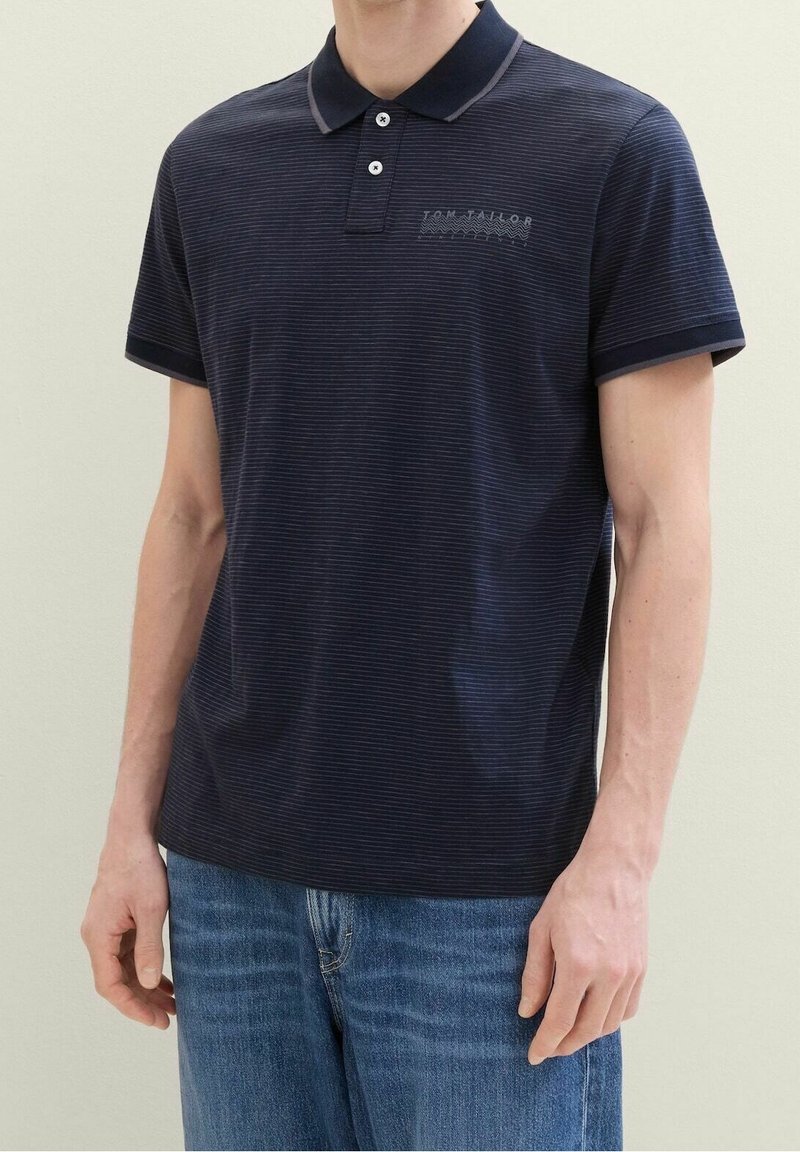 Homme portant un polo bleu foncé à fines rayures avec manches courtes et un jean bleu, debout les bras détendus le long du corps.