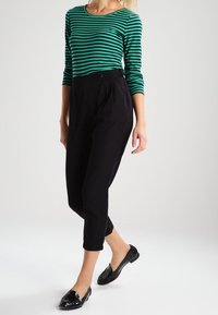 Ein grün und schwarz gestreiftes Langarmoberteil, kombiniert mit hochtaillierten schwarzen Hosen, getragen mit schwarzen Ballerinas, zeigt eine figurbetonte Silhouette.