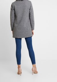 Harmaa pitkä takki, sileä pinta, yhdistetty sinisiin kapeisiin farkkuihin ja nude-sävyisiin nilkkaremmi-korkokenkiin. Korostettu kerrospukeutuminen ja kontrastivärit.