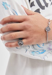 Anello e bracciale in argento con medaglioni circolari caratterizzati da disegni intricati. La mano poggia su un tessuto bianco con motivi blu e grigi.