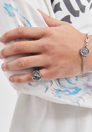 Bague et bracelet en argent ornés de médaillons circulaires avec des designs complexes. Une main repose sur un tissu blanc aux motifs bleus et gris.