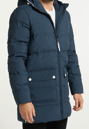 Homme portant une veste d'hiver matelassée bleu marine avec fermeture éclair à l'avant, grandes poches à rabat avec boutons pression, et une capuche.