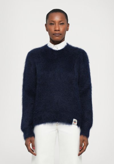 Marineblå, blød strikket sweater med rund halsudskæring og lange ærmer, med en stoffetiket i den nederste kant og båret over hvide bukser.