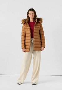 Giacca imbottita marrone con cappuccio foderato in pelliccia, chiusura frontale con zip e design trapuntato orizzontale. Indossata sopra una maglia rossa e pantaloni beige a gamba larga.