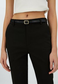Černé kalhoty s hladkým povrchem a předními kapsami, doplněné černým opaskem se zlatou sponou, stylizované s bílým crop topem.