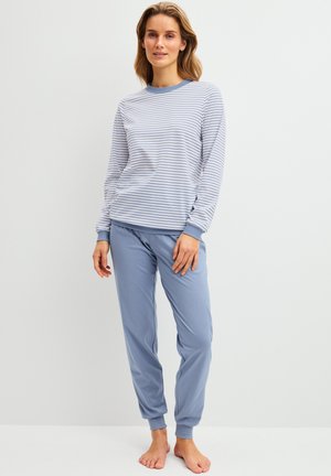 Vrouw staat op blote voeten, gekleed in een witblauw gestreept shirt met lange mouwen en blauwe manchetten, en blauwe losse broek, tegen een effen achtergrond.