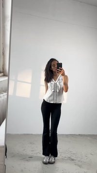 Top branco sem mangas com design frontal em babados, calças ajustadas pretas e ténis cinzentos. Superfície espelhada reflexiva, fundo brilhante.