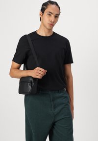 Sort læder crossover taske med en lynlåslukket frontlomme, rektangulær form og justerbar rem, parret med en sort T-shirt og grønne kordfløjl bukser.