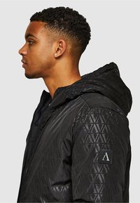 Czarna kurtka z kapturem z geometrycznym wzorem, teksturowanym materiałem, zamkiem błyskawicznym oraz naszywką z logo na lewym rękawie. Gładka powierzchnia, matowe wykończenie.