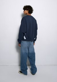 Granatowa bluza z luźnym krojem, z białymi akcentami po bokach. W zestawieniu z luźnymi, jasnobłękitnymi dżinsami i czarnymi trampkami.