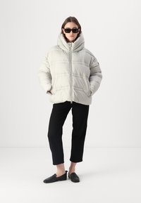 Pufferjack in lichtgrijs materiaal met een hoge kraag en zichtbare stiksels. Gecombineerd met zwarte cropped broeken en zwarte loafers.