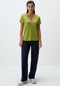 Zielona koszulka z dekoltem w serek i krótkimi rękawami, zestawiona z ciemnoniebieskimi dżinsami o prostym kroju. Gładka tekstura tkaniny; luźny krój. Na nogach białe trampki.
