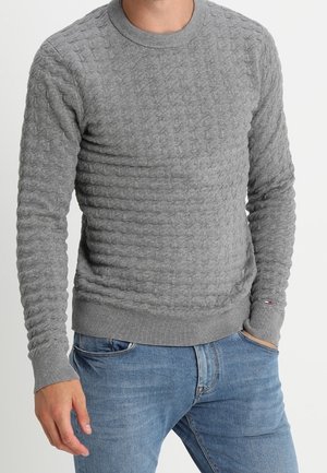 Grauer gestrickter Pullover mit einem strukturierten Muster, Rundhalsausschnitt und gerippten Bündchen. Kombiniert mit blauen Jeans, die einen lässigen Look zeigen.