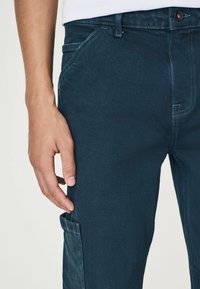 Donkerblauwe broek gemaakt van een geweven stof, met contrasterende stiknaden, een knoopsluiting en zijzakken voor extra functionaliteit.