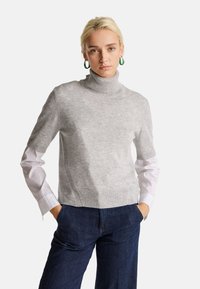 Mladá žena se blond vlasy má na sobě šedý krátký rolák přes bílou dlouhou košili a tmavě modré džíny, s rukama v kapsách.