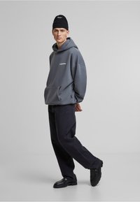 Grijze oversized hoodie met een voorkant zak, witte logo-details, gecombineerd met donkere broeken en zwarte chunky schoenen. Tegen een neutrale achtergrond.