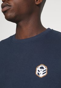 Närbild av en person som bär en marinblå skjorta med en liten vit och orange hexagonal logotyp på bröstet.