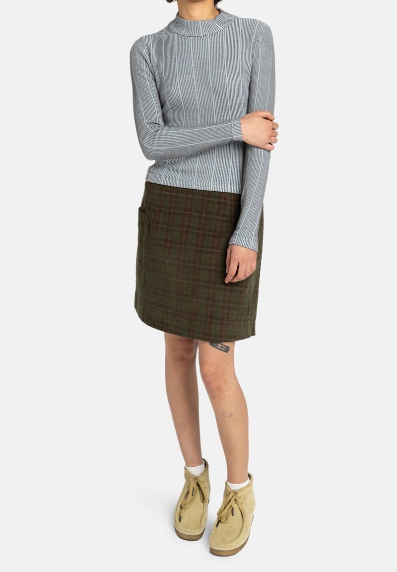 Personne vêtue d'un haut gris côtelé à manches longues, d'une jupe verte à carreaux avec poches, de chaussettes blanches et de bottines beiges à lacets, debout sur un fond blanc.
