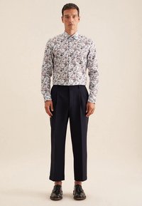 Camicia bianca con motivo floreale, maniche lunghe e colletto a punta, abbinata a pantaloni blu navy scuro e scarpe nere.