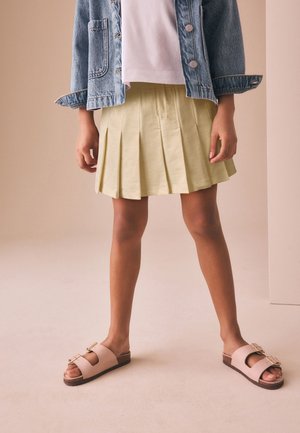 Kind draagt een lichtgroene geplooide rok, wit overhemd, blauwe spijkerjas en beige sandalen met gesp, staand met de benen iets uit elkaar.