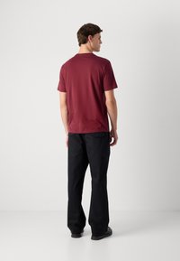 Man in een bordeauxrood T-shirt met korte mouwen en losse zwarte broek, staand met de rug naar de camera tegen een effen lichte achtergrond.