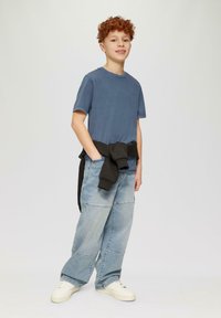 Blauwe T-shirt met korte mouwen gecombineerd met lichtblauwe, losse patchwork denim jeans. Zwart vest om de taille geknoopt, witte sneakers.