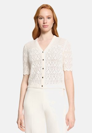 Rødhåret kvinde iført en hvid, kortærmet, hæklede cardigan med knapper og geometriske mønstre samt hvide bukser med høj talje.