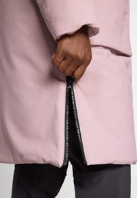 Ruka držící a částečně rozepínající černý zip na světle růžovém kabátu přes černé kalhoty.