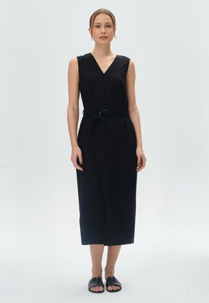 Schwarzes, ärmelloses Kleid mit V-Ausschnitt und tailliertem Bund. Gerader Schnitt, knielang, aus glattem Stoff. Getragen mit schwarzen flachen Sandalen.