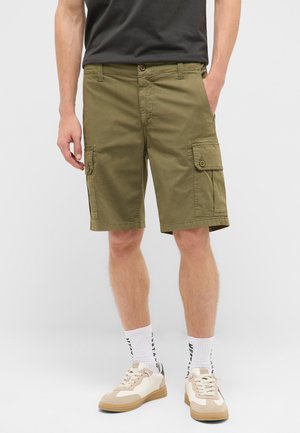 Mann trägt olivgrüne Cargoshorts, weiße Socken mit schwarzem Text, beige und weiße Sneakers, steht mit einer Hand in der Tasche.
