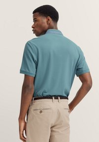 Polohemd aus tealfarbenem Stoff mit einem blauen Kragen, kurzen Ärmeln und einer legeren Passform. Kombiniert mit beigefarbenen Hosen und einem geflochtenen schwarzen Gürtel.
