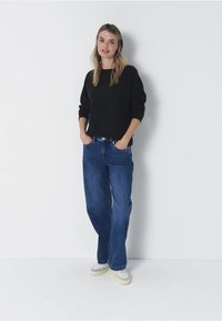 Pull noir à manches longues associé à un jean bleu. Le jean a une coupe décontractée et un léger délavage. Le modèle porte des sneakers de couleur claire.